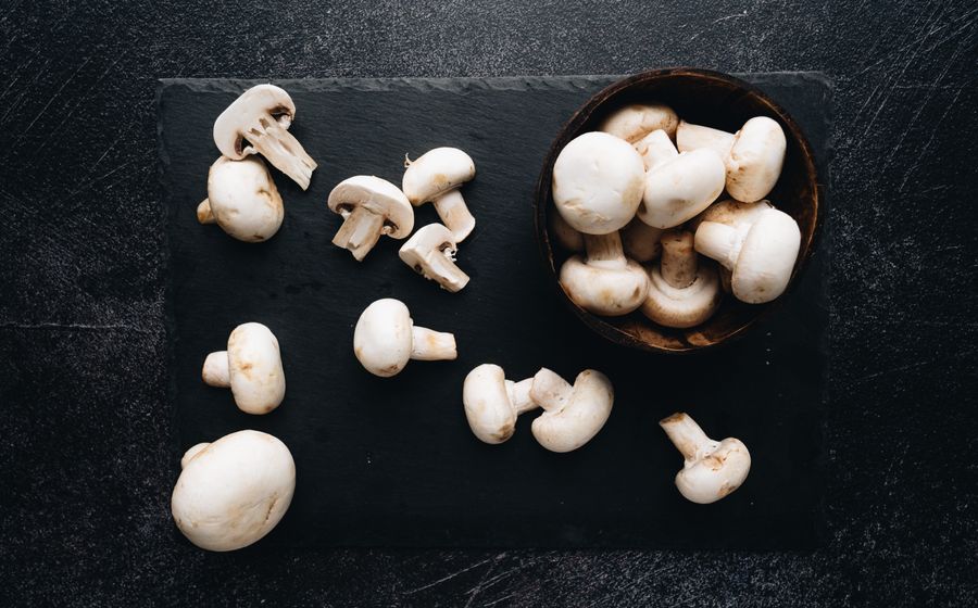 Des champignons de Paris blancs et frais, entiers ou coupés en deux, posés sur une ardoise noire à côté d'un bol en bois rempli de champignons, vus du dessus