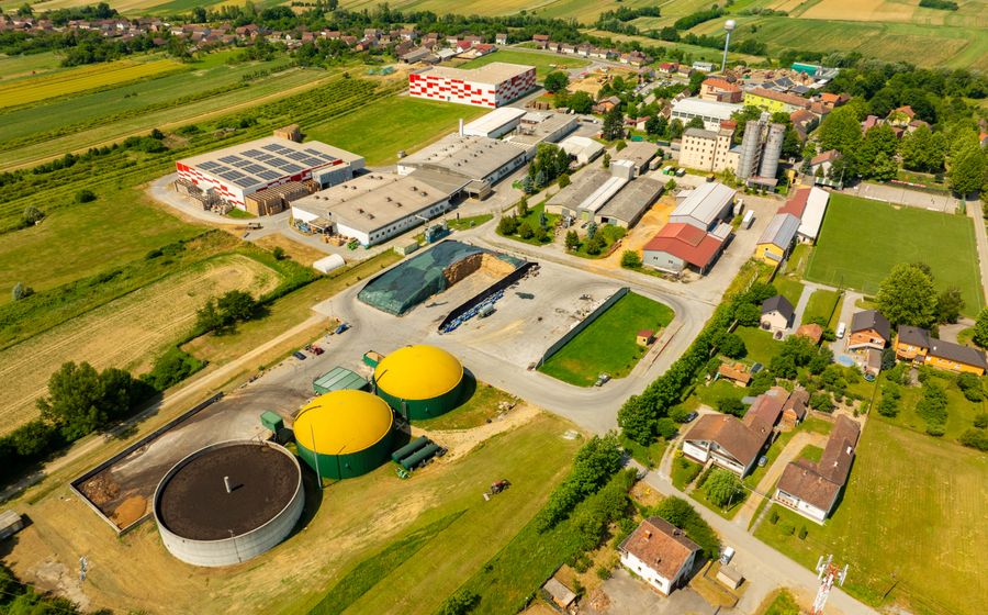 Vue aérienne d'une unité de méthanisation en zone rurale, avec deux digesteurs coiffés de dômes jaunes, une fosse ronde de stockage et un silo de fourrage bâché, entourés de champs et de maisons